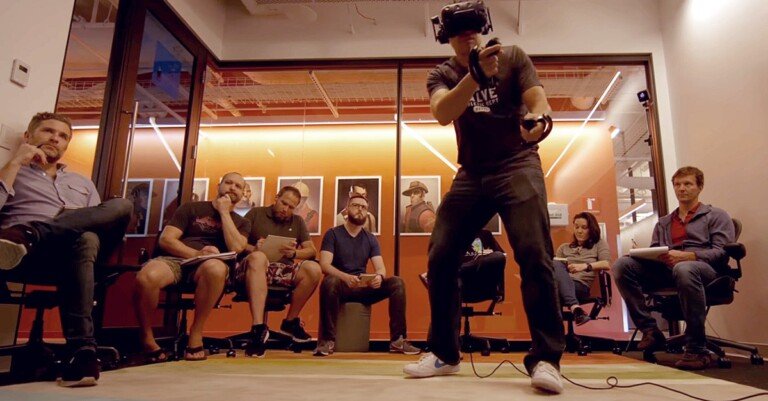 Un uomo indossa un visore VR e usa i controller mentre altre persone lo osservano in un ufficio.